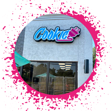 northport cookie plug cookie bakery in tuscaloosa al cookie delivery gray stone storefront with a blue, purple, and pink sign in graffiti type that says 'Cookie' with an electrical plug attached to it with cookie baking racks in the window.
